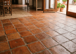 Room interior with terracotta tile floor, wooden table, chairs, and open doors leading to a garden with potted plants